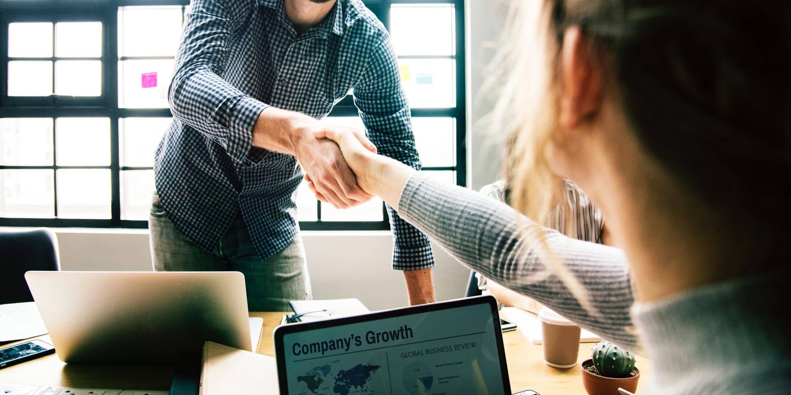A business meeting scene with two people shaking hands, a laptop displaying a company's growth chart, and office items on a table.